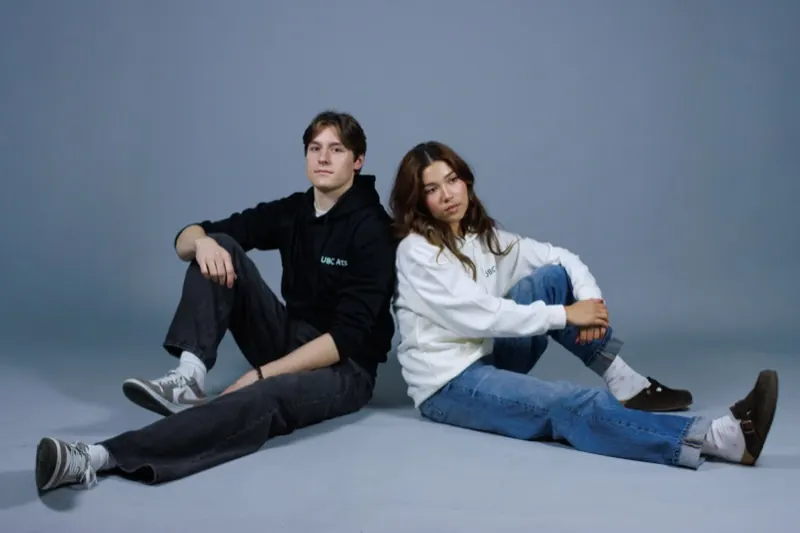 Two models sitting on the studio floor in black and cream hoodies with 'UBC Arts' on the chest