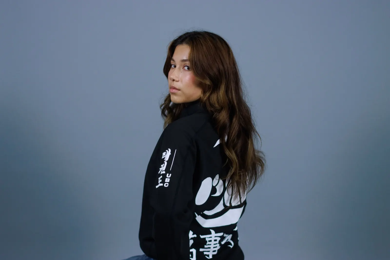 Model in a black custom team jacket with a large white back print, against a blue-grey studio backdrop
